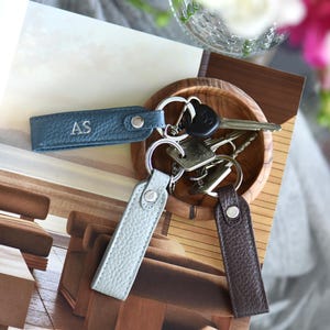 Op de afbeelding: Leren sleutelhangers in blauw, lichtblauw en bruin, elk met een zilveren sleutelring. E&eacute;n sleutelhanger is voorzien van de letters "AS". De sleutelhangers zijn gerangschikt met sleutels in een houten kom.