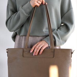May include: A large, taupe-colored leather tote bag with long handles. The bag has a rectangular shape and a textured surface. The person in the image is wearing a green sweatshirt and holding the bag.