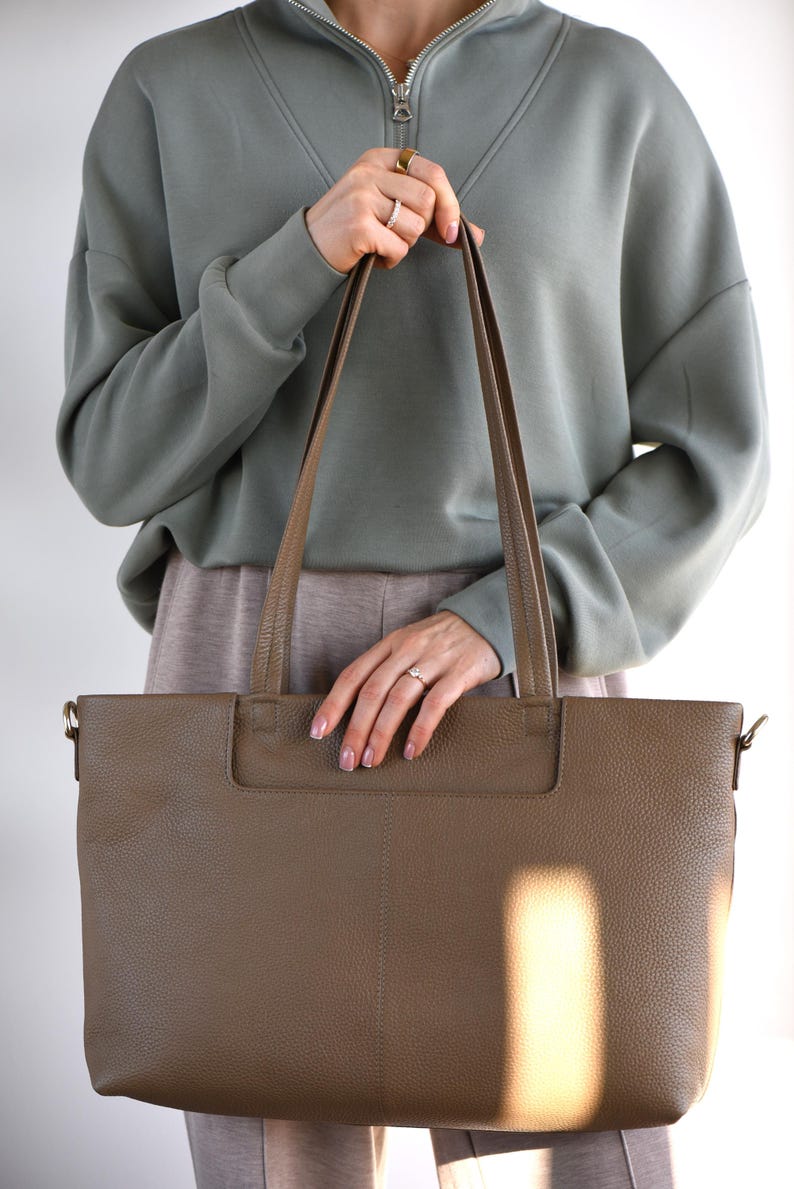 May include: A large, taupe-colored leather tote bag with long handles. The bag has a rectangular shape and a textured surface. The person in the image is wearing a green sweatshirt and holding the bag.