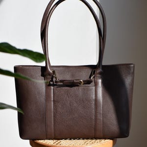 May include: A brown leather tote bag with a rectangular shape and two long handles. The bag features a small front pocket with a decorative buckle and is sitting on a wooden stool.