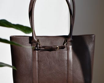 May include: A brown leather tote bag with a rectangular shape and two long handles. The bag features a small front pocket with a decorative buckle and is sitting on a wooden stool.