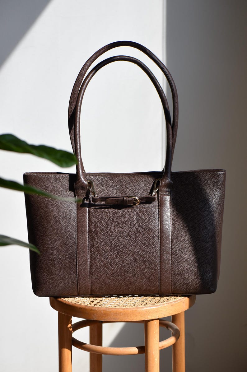May include: A brown leather tote bag with a rectangular shape and two long handles. The bag features a small front pocket with a decorative buckle and is sitting on a wooden stool.