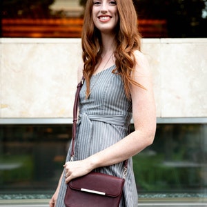 May include: A woman wearing a gray and white striped dress with a burgundy crossbody bag. The bag has a gold strap and a gold accent on the front.