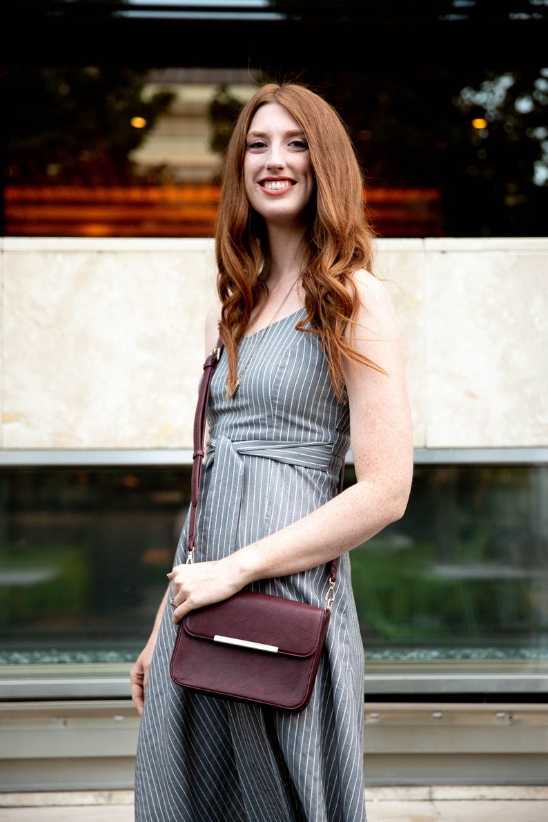 May include: A woman wearing a gray and white striped dress with a burgundy crossbody bag. The bag has a gold strap and a gold accent on the front.