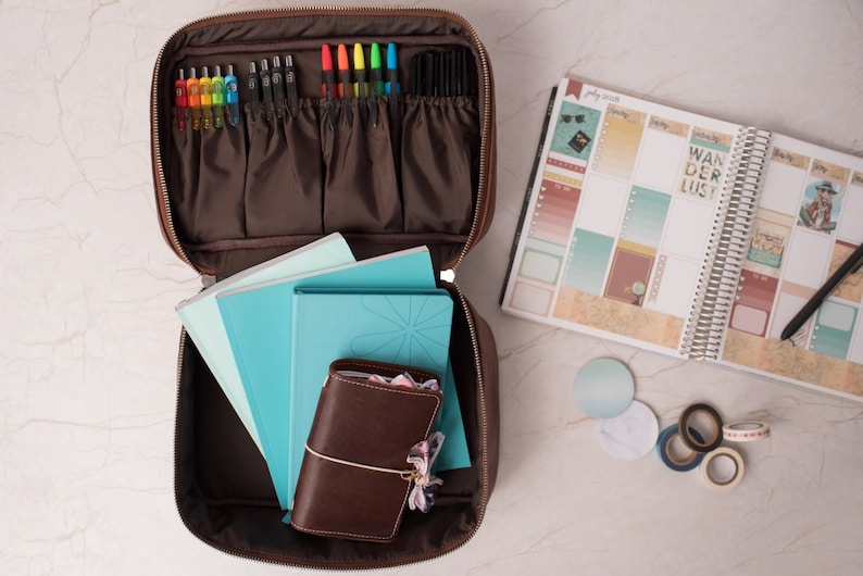 Puede incluir: Un estuche organizador marr&oacute;n con cremallera que contiene varios bol&iacute;grafos coloridos en bucles el&aacute;sticos y varios cuadernos de color verde azulado. Un peque&ntilde;o diario encuadernado en cuero marr&oacute;n se encuentra frente a los cuadernos. Un planificador abierto con el texto WANDERLUST.