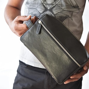 May include: Black leather rectangular bag with a silver zipper closure. The bag has a handle on one side and is held by a person. The bag appears to be new and unused.