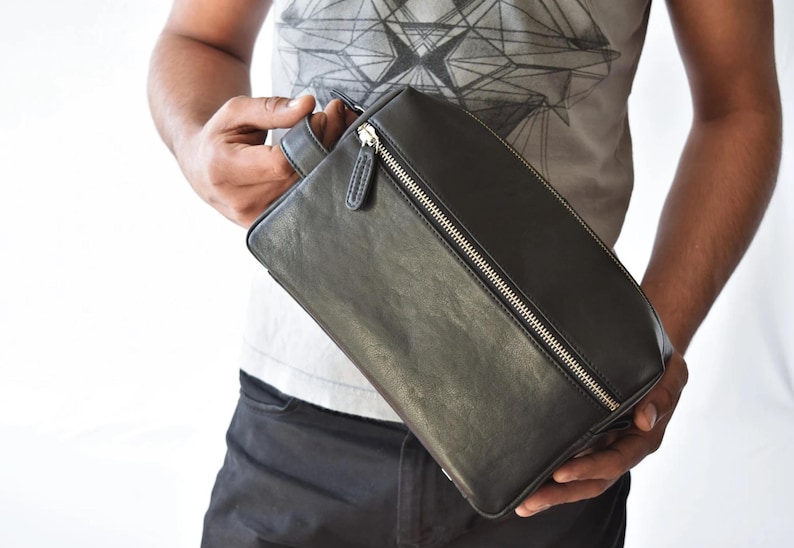 May include: Black leather rectangular bag with a silver zipper closure. The bag has a handle on one side and is held by a person. The bag appears to be new and unused.
