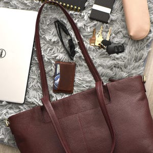 May include: A burgundy leather tote bag with long handles, a laptop, wallet, keys, glasses, and a notebook. The items are arranged on a gray, textured surface. A pink glasses case and a black card holder are also visible.