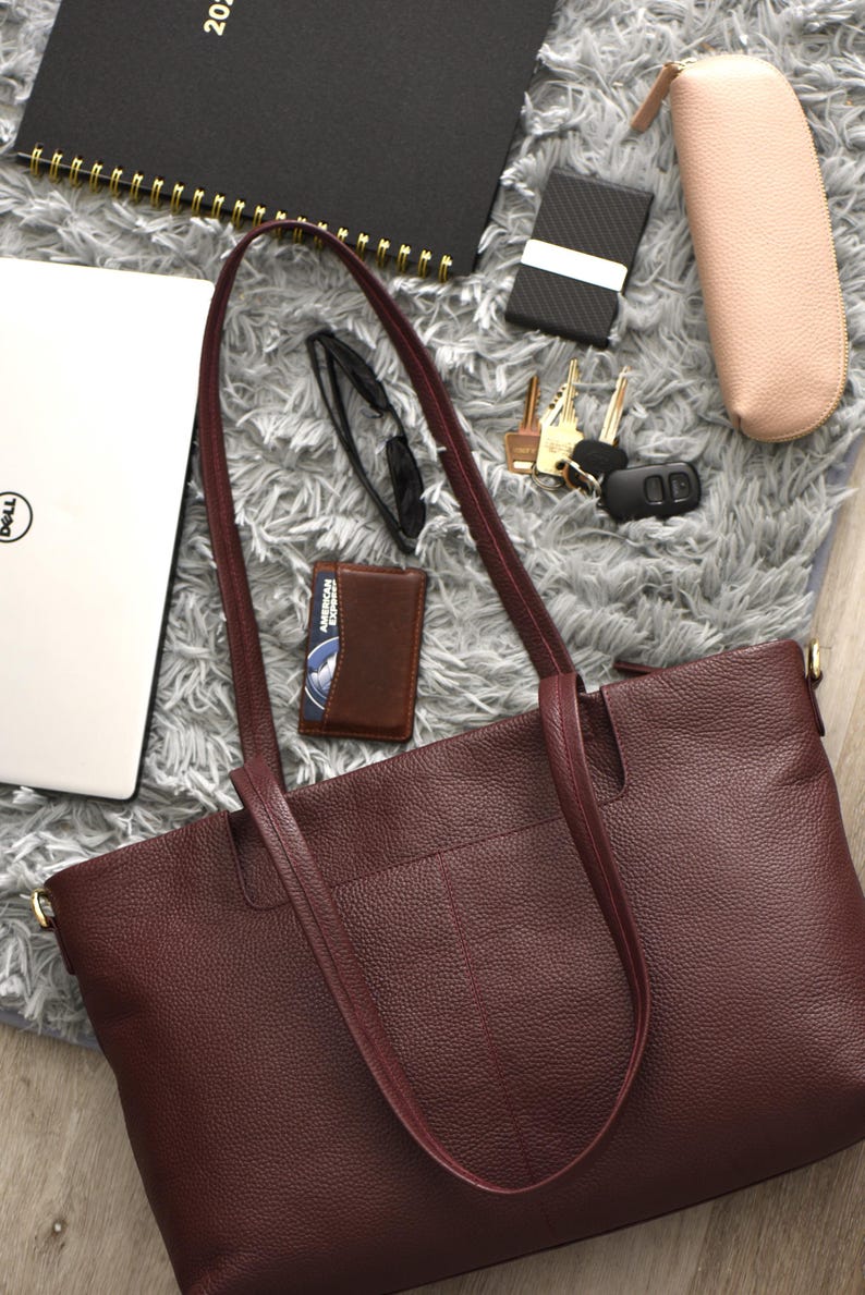 May include: A burgundy leather tote bag with long handles, a laptop, wallet, keys, glasses, and a notebook. The items are arranged on a gray, textured surface. A pink glasses case and a black card holder are also visible.