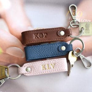 Op de afbeelding: Drie leren sleutelhangers in bruin, blauw en roze, elk met een zilveren sleutelring en een drukknoopsluiting. De bruine sleutelhanger heeft de initialen "KOP" in reli&euml;f, de roze sleutelhanger heeft "KLV", en de blauwe sleutelhanger is leeg. Sleutels zijn bevestigd.