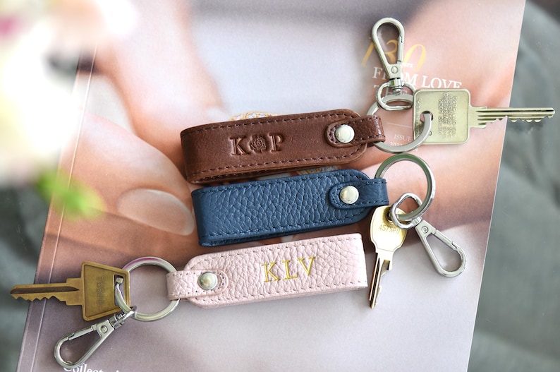 Op de afbeelding: Drie leren sleutelhangers in bruin, blauw en roze, elk met een zilveren sleutelring en een drukknoopsluiting. De bruine sleutelhanger heeft de initialen "KOP" in reli&euml;f, de roze sleutelhanger heeft "KLV", en de blauwe sleutelhanger is leeg. Sleutels zijn bevestigd.
