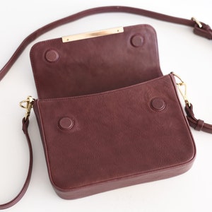 May include: A burgundy leather crossbody bag with a gold metal closure and a long adjustable strap.