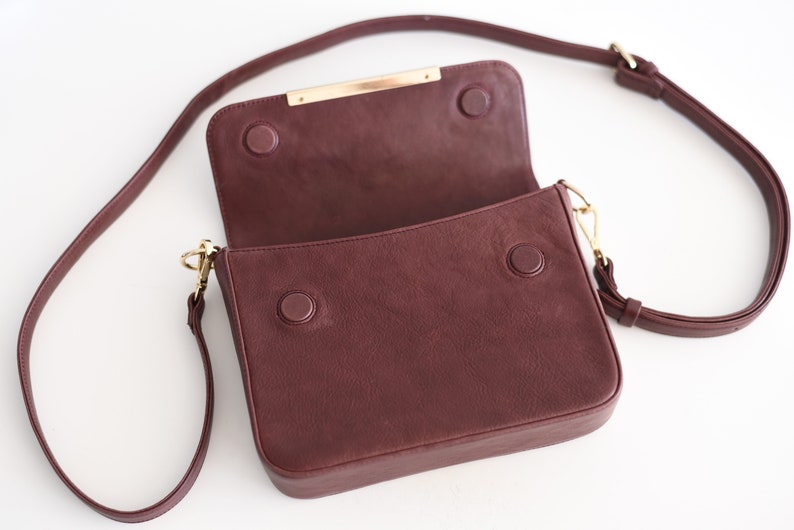 May include: A burgundy leather crossbody bag with a gold metal closure and a long adjustable strap.
