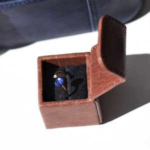 May include: A small brown leather ring box with the lid open, revealing a gold ring with a blue gemstone.