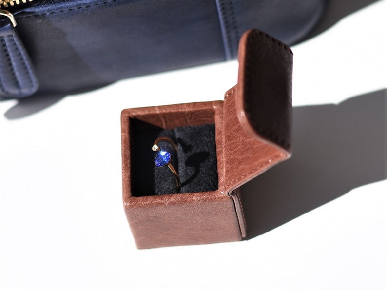 May include: A small brown leather ring box with the lid open, revealing a gold ring with a blue gemstone.