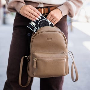 May include: A tan pebbled leather backpack with a front zipped pocket and gold-tone hardware. The backpack features the embossed letters WOF on the upper front panel. A black and white instant camera is being placed inside the main compartment.