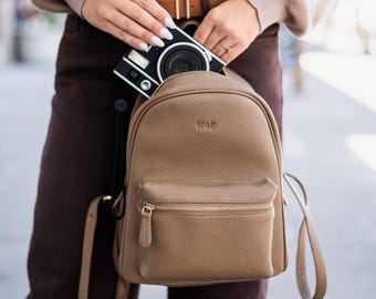 May include: A tan pebbled leather backpack with a front zippered pocket and gold-tone hardware. The backpack features the embossed letters WOF on the upper front panel. A black and white instant camera is being placed inside the main compartment.