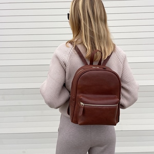 May include: A brown leather backpack with a front zippered pocket and top handle. The backpack has adjustable shoulder straps and a classic design. The bag is worn by a person wearing a light pink sweater and gray sweatpants.