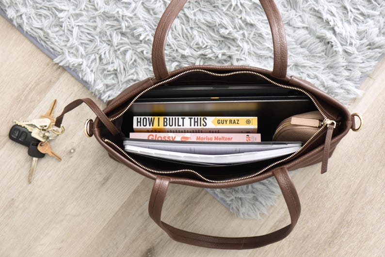 May include: A brown leather tote bag with a zipper closure, filled with books, a laptop, and a small pouch. The bag has two handles and a gold-colored zipper. Keys are visible next to the bag.