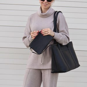 May include: A woman wearing a beige turtleneck sweater and matching pants, holding a black tote bag and a black clutch with a silver chain. She is also wearing black sunglasses. The outfit is neutral-toned and appears to be made of a soft material.