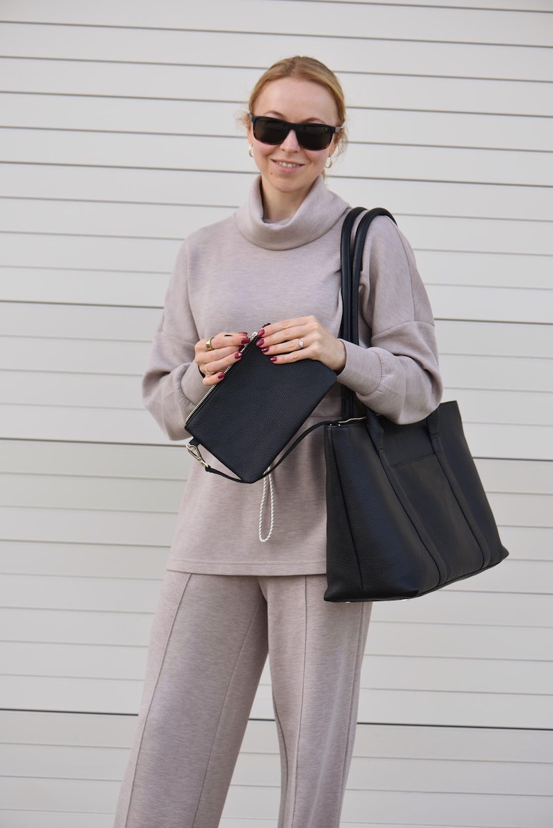 May include: A woman wearing a beige turtleneck sweater and matching pants, holding a black tote bag and a black clutch with a silver chain. She is also wearing black sunglasses. The outfit is neutral-toned and appears to be made of a soft material.
