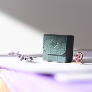 May include: A small teal green ring box with a heart embossed on the lid. The box is sitting on a white surface next to a sprig of lavender and a gold ring with pink gemstones.