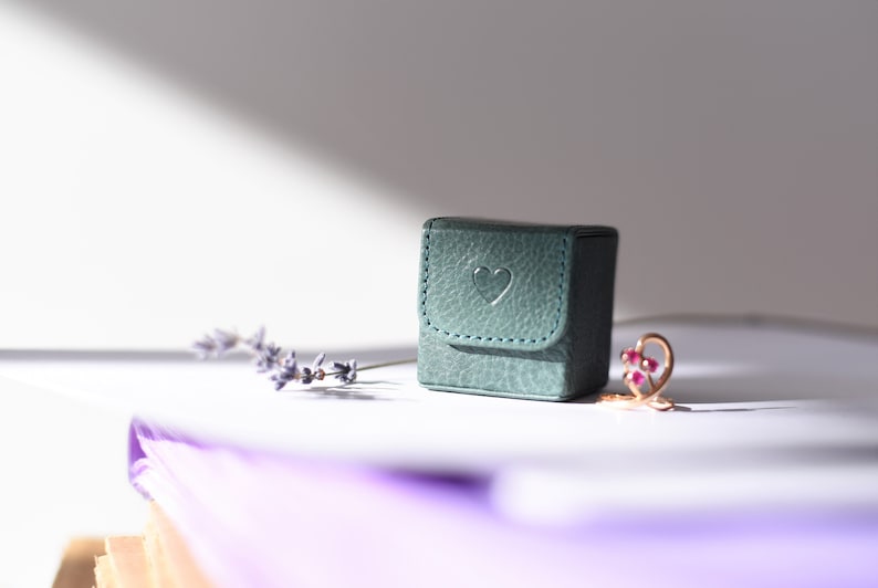 May include: A small teal green ring box with a heart embossed on the lid. The box is sitting on a white surface next to a sprig of lavender and a gold ring with pink gemstones.
