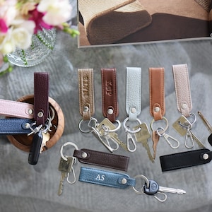 Op de afbeelding: Leren sleutelhangers in diverse kleuren, waaronder roze, blauw, bruin en zwart. Sommige sleutelhangers hebben initialen in reli&euml;f. Elke sleutelhanger is bevestigd aan een metalen ring met sleutels.