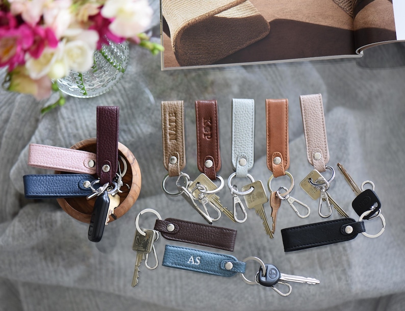 Op de afbeelding: Leren sleutelhangers in diverse kleuren, waaronder roze, blauw, bruin en zwart. Sommige sleutelhangers hebben initialen in reli&euml;f. Elke sleutelhanger is bevestigd aan een metalen ring met sleutels.