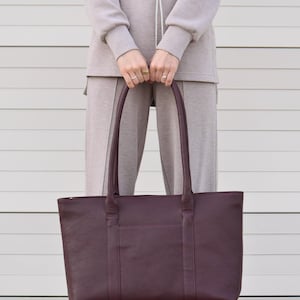 May include: A burgundy leather tote bag with two handles and a rectangular shape. The bag has a textured surface and a pocket on the front. The person is wearing a beige sweatsuit.