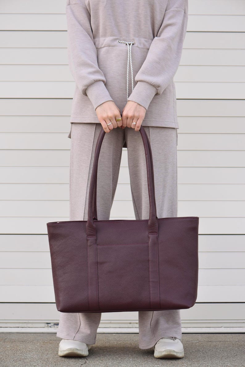 May include: A burgundy leather tote bag with two handles and a rectangular shape. The bag has a textured surface and a pocket on the front. The person is wearing a beige sweatsuit.