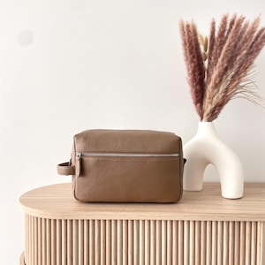 May include: A brown leather travel bag with a silver zipper and a small handle. The bag sits on a light wood table next to a white vase with dried pink grass. The table has a ribbed design.