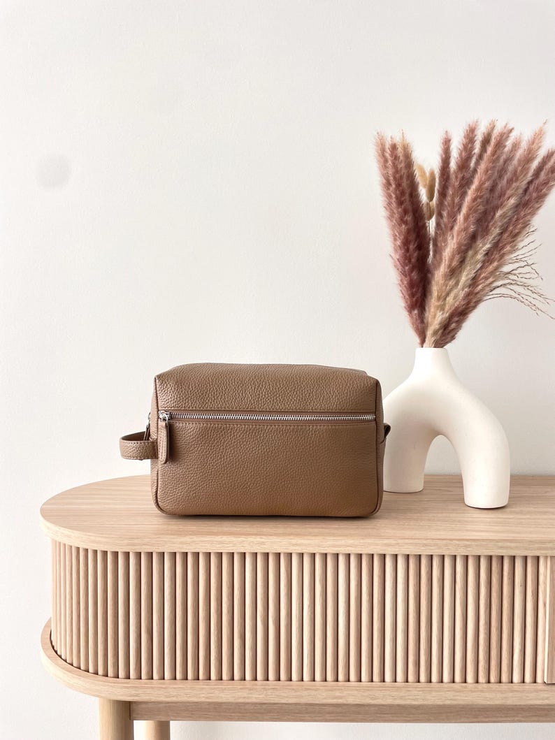 May include: A brown leather travel bag with a silver zipper and a small handle. The bag sits on a light wood table next to a white vase with dried pink grass. The table has a ribbed design.