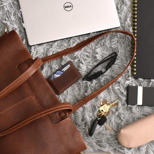 May include: A brown leather tote bag with a matching wallet, keys, sunglasses, and a laptop. A black notebook with "2026" printed on it, a card holder, and a pink glasses case are also visible. The items are arranged on a gray, textured surface.