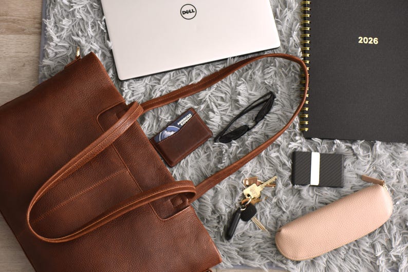 May include: A brown leather tote bag with a matching wallet, keys, sunglasses, and a laptop. A black notebook with "2026" printed on it, a card holder, and a pink glasses case are also visible. The items are arranged on a gray, textured surface.