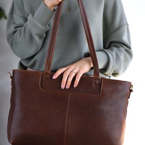 May include: A brown leather tote bag with long handles. The bag has a rectangular shape with a textured surface and a pocket on the front. The handles are a darker brown than the bag itself. The bag is held by a person wearing a green sweatshirt.