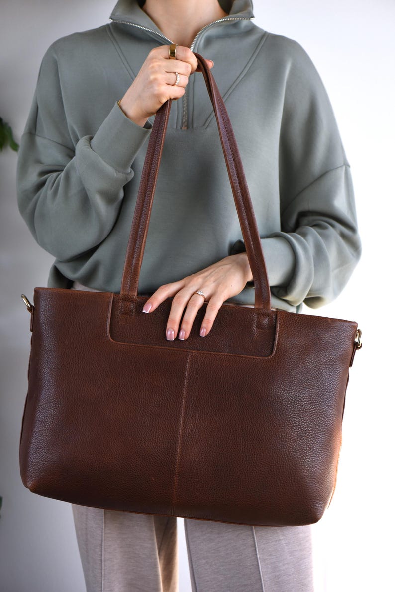 May include: A brown leather tote bag with long handles. The bag has a rectangular shape with a textured surface and a pocket on the front. The handles are a darker brown than the bag itself. The bag is held by a person wearing a green sweatshirt.