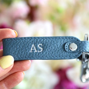 Op de afbeelding: Een blauwe leren sleutelhanger met de geborduurde initialen "AS". De sleutelhanger heeft een zilverkleurige drukknoopsluiting en een metalen sleutelring met sleutels. De achtergrond is een zachte focus van roze en witte bloemen.