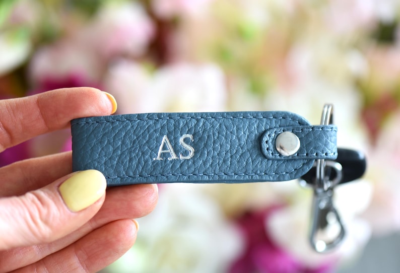 Op de afbeelding: Een blauwe leren sleutelhanger met de geborduurde initialen "AS". De sleutelhanger heeft een zilverkleurige drukknoopsluiting en een metalen sleutelring met sleutels. De achtergrond is een zachte focus van roze en witte bloemen.