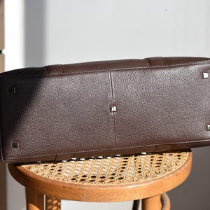 May include: A dark brown leather tote bag with a textured surface. The bag has a rectangular shape with metal feet on the bottom. The bag is sitting on a round wooden stool.