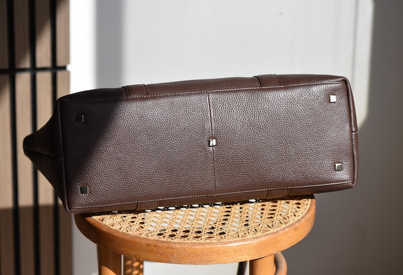 May include: A dark brown leather tote bag with a textured surface. The bag has a rectangular shape with metal feet on the bottom. The bag is sitting on a round wooden stool.