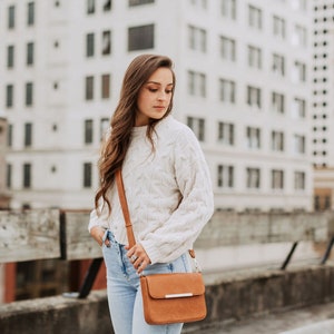 May include: A woman wearing a white cable knit sweater and blue jeans is carrying a brown leather crossbody bag with a silver buckle.