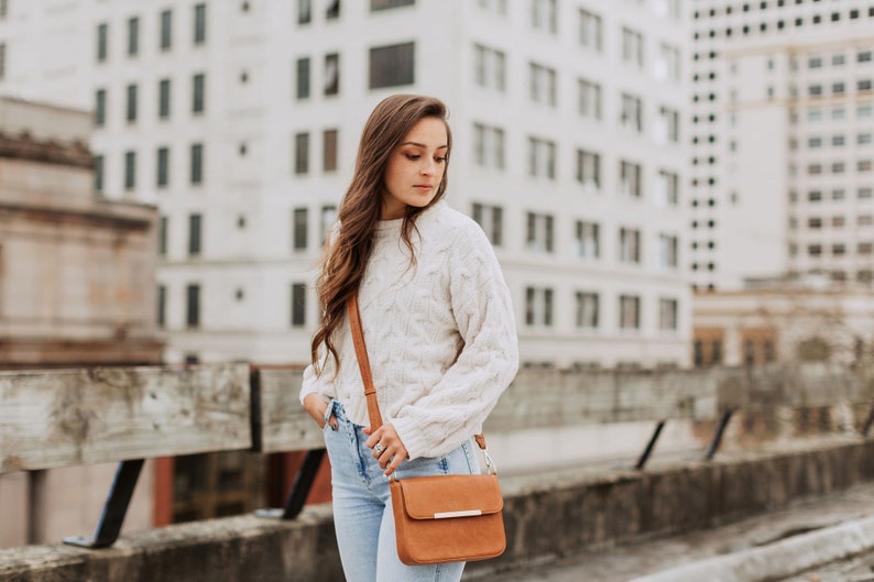 May include: A woman wearing a white cable knit sweater and blue jeans is carrying a brown leather crossbody bag with a silver buckle.
