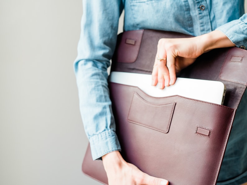 May include: A burgundy leather laptop sleeve with a zipper closure and a front pocket. The sleeve is open and a laptop is partially visible inside.