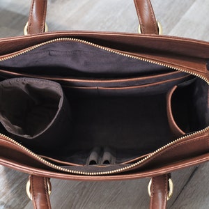 The inside of a brown leather handbag with a zipper closure. The bag has multiple pockets and compartments, including a large main compartment, two pen loops, four smaller pockets, a large zippered pocket, a water bottle holder, and a small charger pocket. The bag is lined with a dark brown fabric.