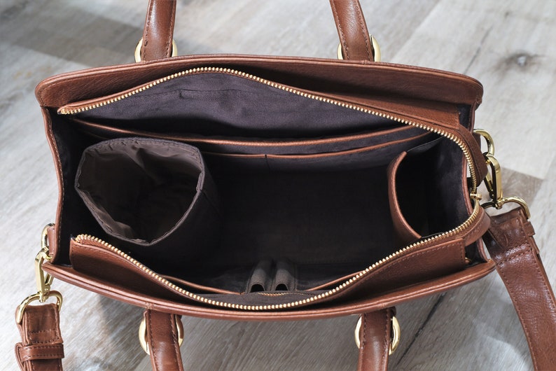 The inside of a brown leather handbag with a zipper closure. The bag has multiple pockets and compartments, including a large main compartment, two pen loops, four smaller pockets, a large zippered pocket, a water bottle holder, and a small charger pocket. The bag is lined with a dark brown fabric.