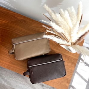 May include: Two leather toiletry bags in the image. One is a light tan color, and the other is a dark brown. Both have a rectangular shape with a zipper closure and a small handle on the side. The bags are on a wooden surface.