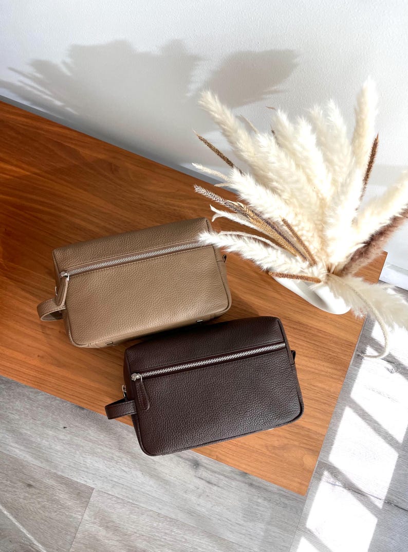 May include: Two leather toiletry bags in the image. One is a light tan color, and the other is a dark brown. Both have a rectangular shape with a zipper closure and a small handle on the side. The bags are on a wooden surface.