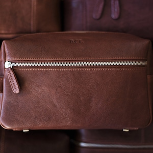 May include: A brown leather toiletry bag with a silver zipper and a small handle. The bag has the initials "RCL" embossed on the front. Other similar bags are visible in the background.