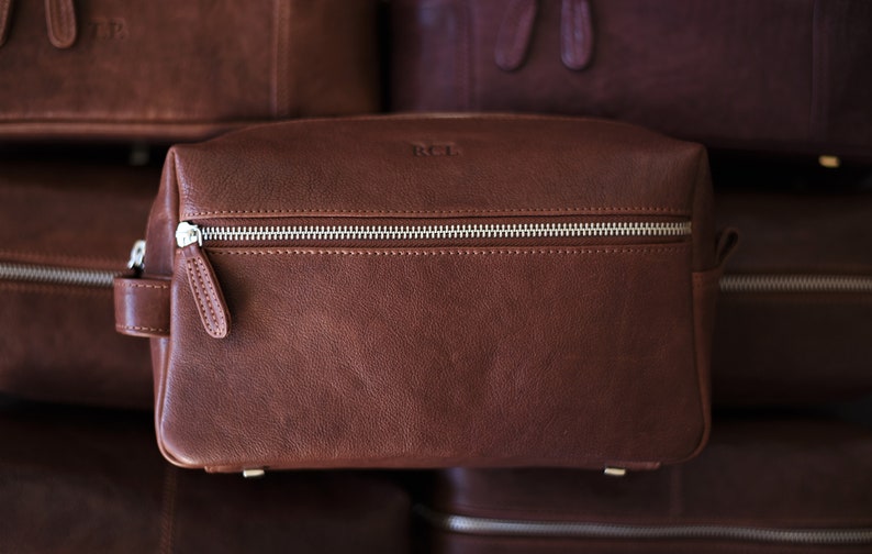 May include: A brown leather toiletry bag with a silver zipper and a small handle. The bag has the initials "RCL" embossed on the front. Other similar bags are visible in the background.